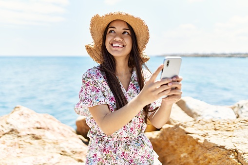 Young latin girl wearing summer hat using smartphone at the beach.