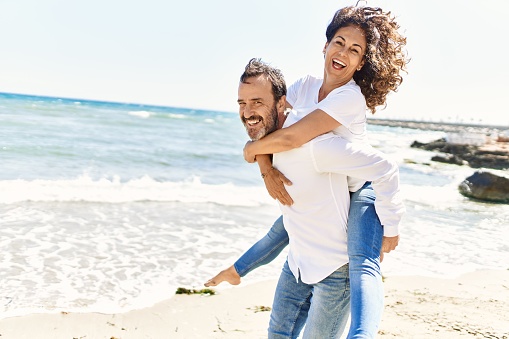 Middle age hispanic man smiling happy holding woman on his back at the beach.