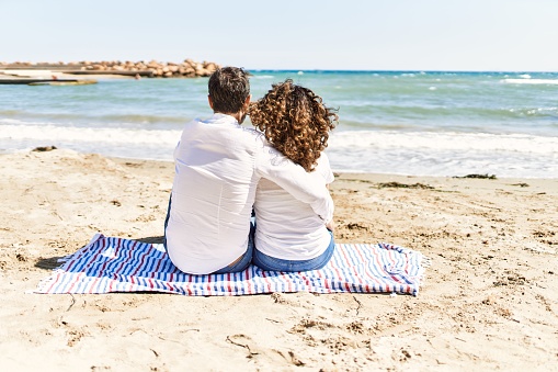 Middle age hispanic couple on back view sitting on the towel hugging at the beach.