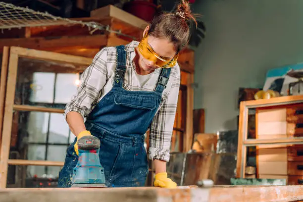 A portrait of a woman wearing protective glasses and gloves working in the workshop using a disc sander. Upcycling concept.