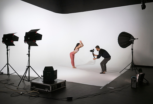 Young beautiful woman wearing sportswear, practicing yoga, while professional photographer shooting and filming in photo studio with lighting equipments