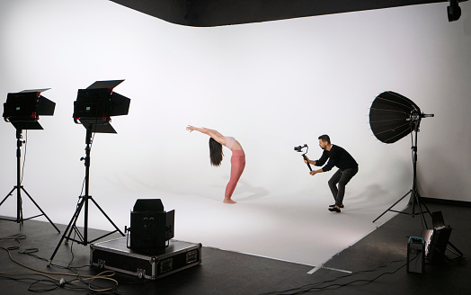 Young beautiful woman wearing sportswear, practicing yoga, while professional photographer shooting and filming in photo studio with lighting equipments