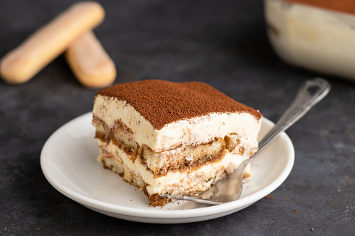 a tilt down shot of a piece of tiramisu with ladyfingers with a dish of tiramisu in the back on a dark background