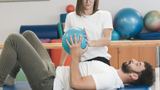 Female physiotherapist and male patient during rehabilitation therapy. Ccore strengthening exercises with pilates ball with therapist assistance.