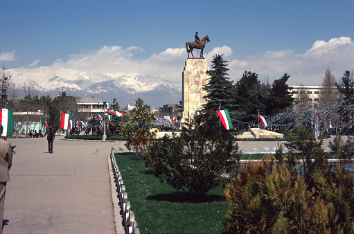 Park Shahr in Tehran, decorated in flags and lights.  There is a statue on a large plinth commemorating a military figure on horseback.  This statue has since been removed and replaced with an abstract sculpture.  In the foreground, in front of the plinth, there is a water feature with fountains.  The Elburz mountains feature in the distance.  Recorded on Kodachrome approximately on the date created, shown.