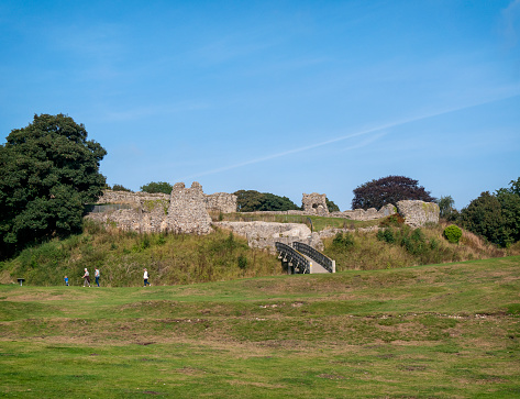 A line of tourists passing part of the ruins of the keep and defences at Castle Acre Castle in the ancient village of Castle Acre in Norfolk, Eastern England, on a sunny day in September. The motte-and-bailey castle was built by order of William de Warenne during the 1070s near the junction of the River Nar and Peddars Way. Very little is left of the castle now but the village also contains the ruins of a Cluniac Priory and an imposing Bailey Gate, so there is much of interest to see in this now small village, which was once a fortified town.