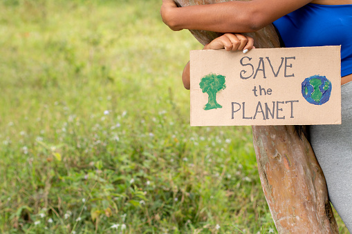 Unknown dark-skinned woman hugging a tree while holding a sign asking to save the planet. Copy space. Care for planet earth, save nature.