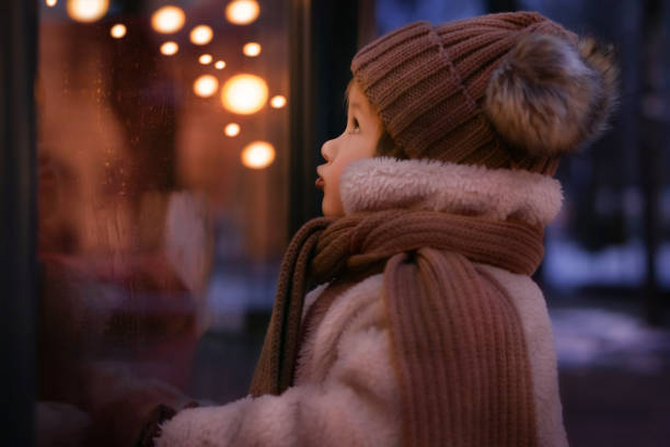 Cute girl is looking to the shop window in Christmas' eve Sweet girl looking into the decorated window. The Christmas' lights reflected in window. window shopping at night stock pictures, royalty-free photos & images