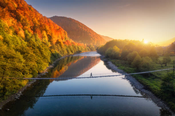 Photo of Aerial view of silhouette of walking man on the suspension bridge, river, mountains, red trees, green grass, orange sun at sunset in autumn. Colorful landscape with forest, lake, reflection in water