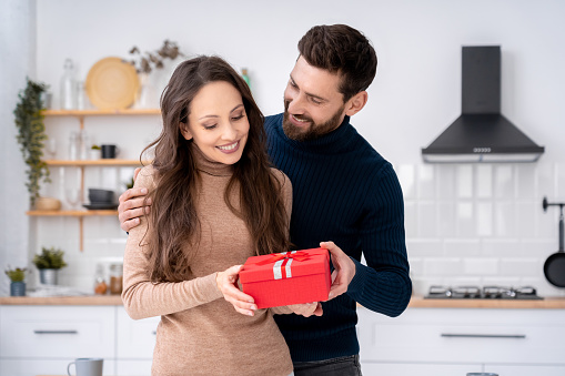 Loving husband surprising charming smiling wife with present gift box on romantic day. Young happy loving family couple celebrating birthday, New Year and Christmas, Valentines day, winter holidays
