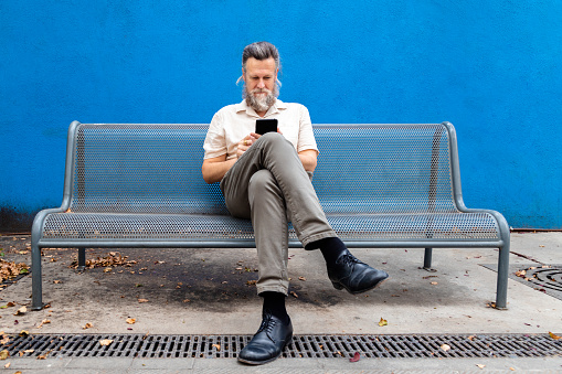 Mature caucasian man sitting on a bench using his mobile phone. Blue background. Copy space. Lifestyle concept.