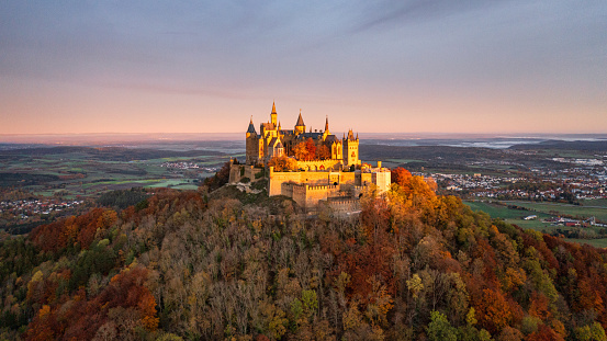 Bisingen, Baden Württemberg, Germany - October, 25th 2020: Majestic spot-lit Hohenzollern Castle - Burg Hohenzollern hilltop castle 16:9 panorama aerial drone point of view  in sunrise light during autumn season. Colorful surrounding autumn colored trees around the hill of Burg Hohenzollern. Swabian Alb, Baden Württemberg, Southwest Germany, Germany, Europe.