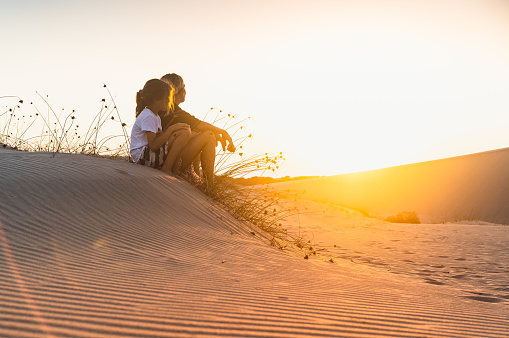 side view portrait of young woman and her child sitting on sand dunes at sunrise and watching the sun going over the horizon