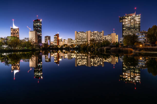 Illuminated Buildings of Business District in the City of Vienna Austria in Twilight with Skyline Reflecting in Water of River Danube at Clear Evening Sky, Wide View