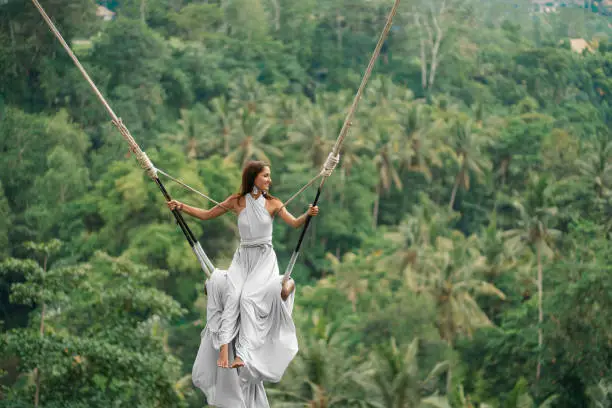 Tanned beautiful woman in a long white dress with a train, riding on a swing. In the background, a rainforest and palm trees. Copy space.