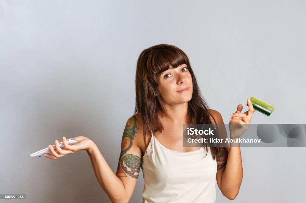 Bank cards and online shopping. A brunette woman holds a Bank card and a mobile phone, shrugging in surprise. Copy space Bank cards and online shopping. A brunette woman holds a Bank card and a mobile phone, shrugging in surprise. Copy space. Refinancing Stock Photo
