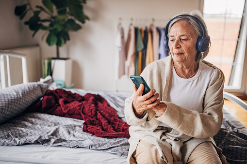 Senior woman sitting on the bed and listening to music on wireless head phones