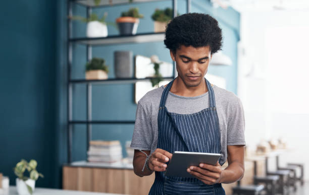 istock Shot of a handsome young waiter standing alone in his cafe and looking contemplative while using a digital tablet 1351999749