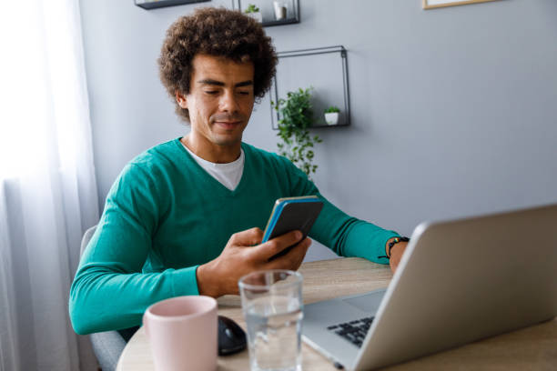 istock Young man sitting at table and reading text messages on smart phone 1352001102