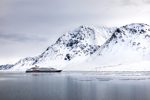 Pristine snow covered mountains of Svalbard with tourist cruise ship in the fiord. Ecotourism is becoming popular in the region, with a number of larger ships now travelling into the Arctic.
