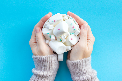 Female hands hold a cup of hot chocolate lavishly decorated with marshmallows on a blue background.