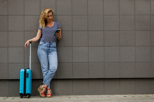 Wide shot of mid adult woman standing by the suitcase she packed, in front of a building on the city street, waiting for a taxi and text messaging a friend via smart phone.