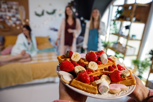 Selective focus shot of unrecognizable woman carrying a nicely arranged plate with fruit, waffles, cookies and marshmallows to serve her to her girlfriends during a slumber party. Focus on foreground.
