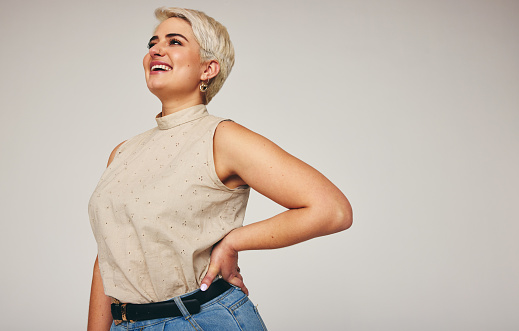 Woman laughing while standing alone against a grey background. Happy mid-adult woman looking away cheerfully in a studio. Fashionable woman wearing denim jeans.