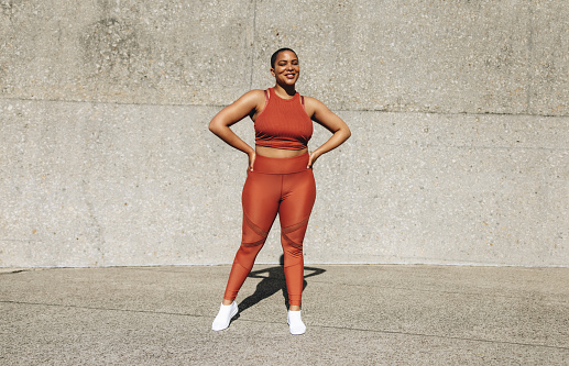 Full length portrait of a happy young sportswoman standing outdoors. Young female athlete smiling at the camera while standing with her hands on her hips.