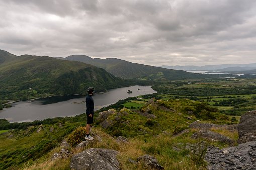 Young cyclist standing on the side of the road looking at the mountains and lake on peninsula Beara Ireland