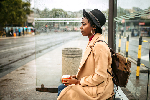Young female black student in rush waiting for a bus at bus station shelter