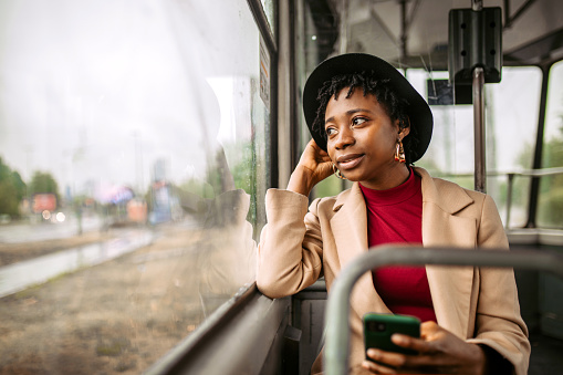 Young smiling woman using smart phone while traveling to work by bus, looking through the window in to rainy cold day