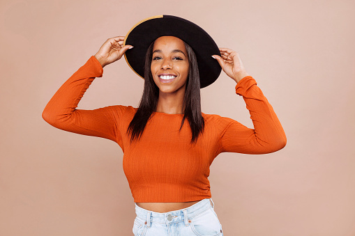 Cheerful mixed race woman with stylish hat and orange shirt, posing in studio, on pastel beige background. Black woman with scar on her forehead with raised arms taking the hat. Close up portrait.