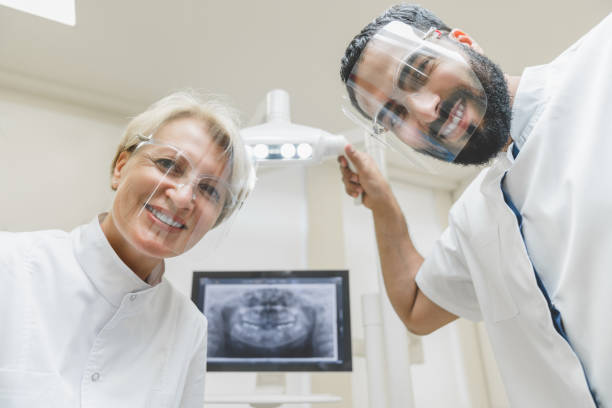 istock Professional smiling two stomatologists assistant orthodontists dentists looking checking up patient`s mouth cavity, curing teeth tooth in protective masks. Low angle view 1352047066