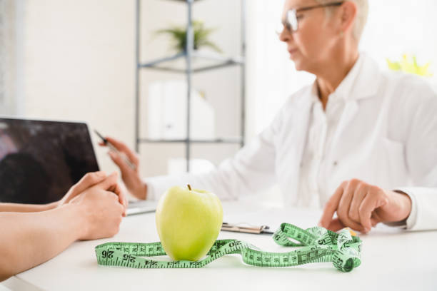 Cropped shot of green apple and measuring tape on dietitian desk. Healthy balanced diet eating concept. Caucasian elderly senior female doctor nutritionist explaining to female patient good nutrition habits. Cropped shot of green apple and measuring tape on dietitian desk. Healthy balanced diet eating concept. Caucasian elderly senior female doctor nutritionist explaining to female patient good nutrition habits. body mass index stock pictures, royalty-free photos & images