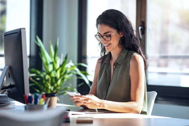 istock Smiling business woman using her mobile phone while working with computer in the office. 1352062917