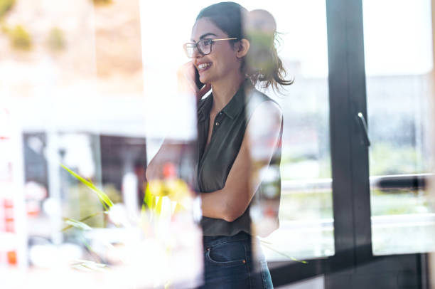 Beautiful woman using her mobile phone while standing next to the window at the office.