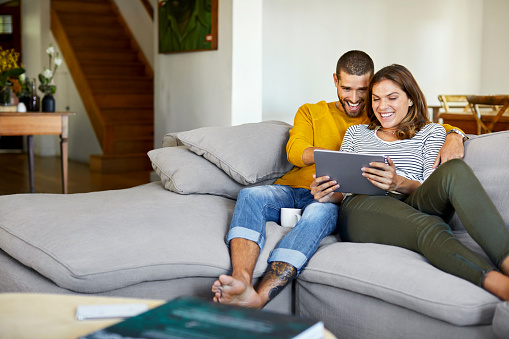 Happy man and woman watching movie on digital tablet. Couple is relaxing on sofa in living room. They are spending leisure time together at home during lockdown.