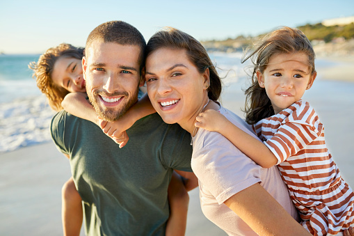 Portrait of smiling mother and father piggybacking children. Boy and girl enjoying with parents at beach. They are on vacation.