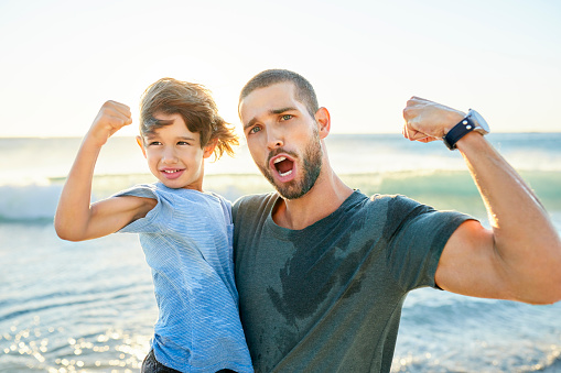 Cheerful father and son flexing muscles at sea shore. Man is enjoying with boy at beach during summer. They are on vacation.