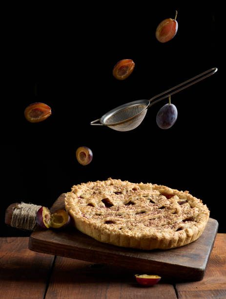 baked round pie with plums on a levitating wooden kitchen board, sieve and livitating slices of plum fruit on a black background