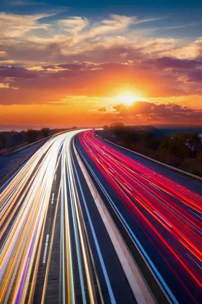 Photo of Trails  of cars lights on the asphalt car road. Sunset time with clouds and sun. Drive forward! Transport creative background. Long exposure, motion and blur.