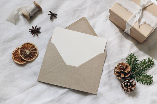 istock Christmas gift wrapping still life. Spruce tree branches, present box and dry orange slices. Blank card mockup, craft envelope and pine cones. White linen tablecloth background. Flatlay, top view. 1352077503
