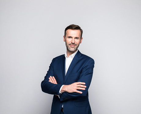 Portrait of mature man wearing navy blue suit and white shirt, standing with arms crossed and looking at camera. Studio shot of male entrepreneur against grey background.