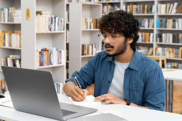 istock Young Indian male student watching webinar using laptop sitting at library 1352080176