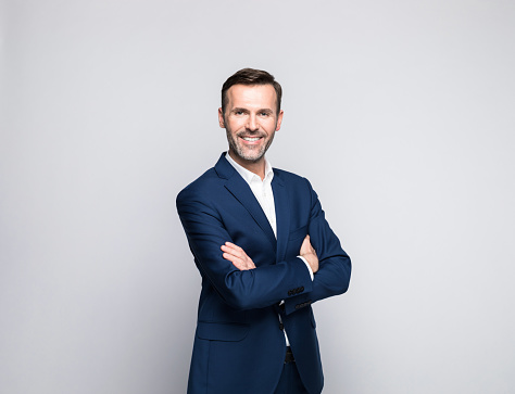 Portrait of mature man wearing navy blue suit and white shirt, standing with arms crossed and smiling at camera. Studio shot of male entrepreneur against grey background.