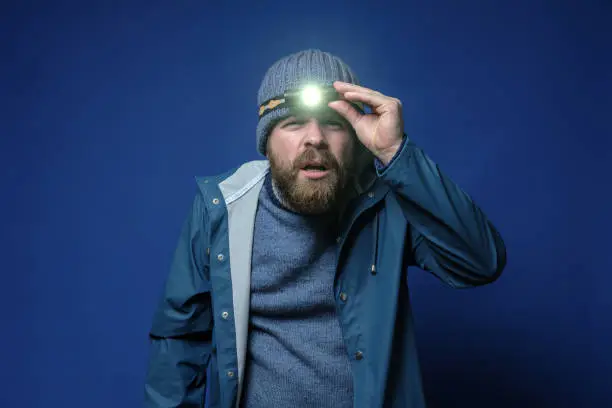 Attractive, bearded man with a headlamp in a knitted hat and a waterproof jacket is trying to see something in the light of a flashlight. Isolated on a blue background.