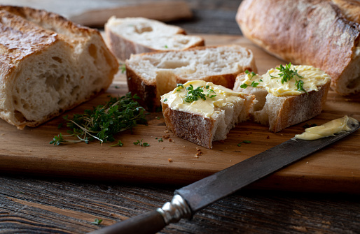 Sliced and fresh french baguette with butter and a rustic table knife served with herbs on a rustic cutting board