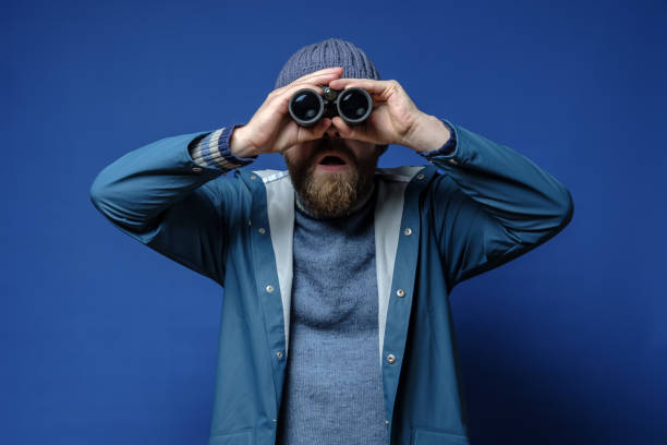 istock Bearded man looks through binoculars, standing on an isolated blue background, with a surprised expression on face and open mouth. 1352082249