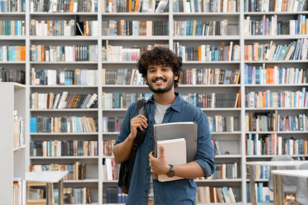 istock Young smiling Indian male student standing in university library with backpack 1352087631